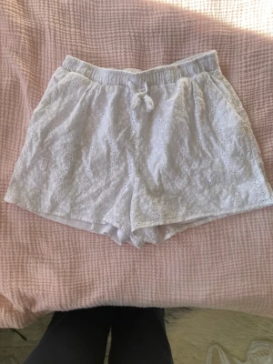 Vita broderade shorts med snörning - Säljer ett par ljusa, vita shorts med elastisk midja och snörning framtill. Shortsen har ett blommigt broderat mönster och är fodrade med ett mjukt innerfoder. Perfekta för varma sommardagar och ger en söt och avslappnad vibe.