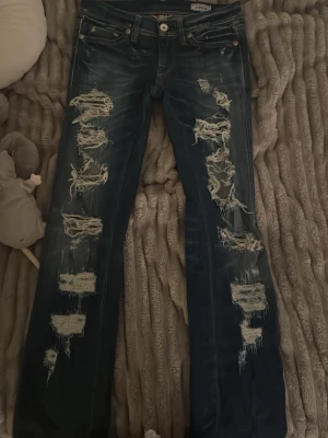 Mörkblå slitna bootcut jeans - Säljer ett par mörkblå bootcut jeans med kraftiga slitningar och hål längs båda benen. Jeansen är lågmidjade. Skriv privat om måtten!!