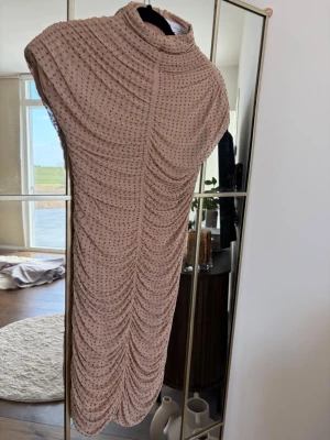 Beige glittrig kortklänning - Supersnygg beige kortklänning med hög hals och glittriga paljettdetaljer. Klänningen har ett rynkat tyg som ger en cool struktur och är ärmlös för en trendig look. Perfekt för dig som vill sticka ut med stilren glamour.