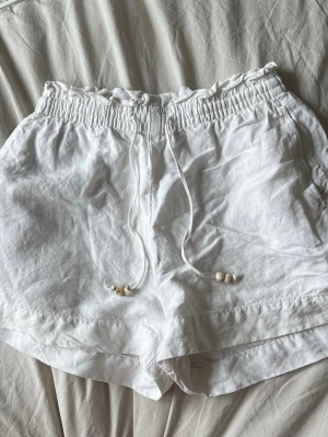 Vita linneshorts med snörning - Säljer ett par vita shorts i linne med elastisk midja och snörning med pärldetaljer. Shortsen har en avslappnad passform och är perfekta för varma dagar. Superlätta och luftiga med enkel design.