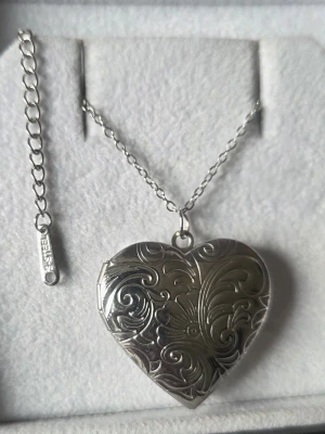 Silverfärgat medaljong hjärthalsband med mönster - Säljer ett silverfärgat hjärthalsband i rostfritt stål. Hänget är ett medaljonghalsband i form av ett hjärta med vackert graverat blommönster och plats för foto inuti. Kedjan är tunn och justerbar. Perfekt accessoar för dig som gillar romantiska detaljer.