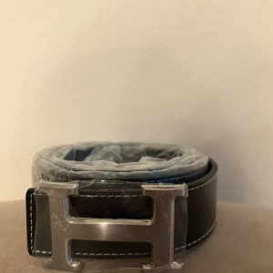 Svart bälte med silver H-spänne - Snyggt svart bälte från med ett stort H-format spänne i silverfärgad metall. Bältet har vita kontrastsömmar längs kanterna och är tillverkat i slätt skinn. Perfekt accessoar för att lyfta vilken outfit som helst.