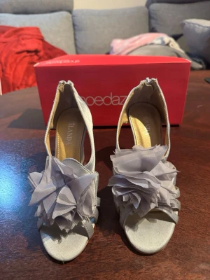 Silvriga kilklackade pumps med blommor - Snygga silvriga pumps från ShoeDazzle med kilklack och öppen tå. Skorna har stora dekorativa blommor i tyg framtill och dragkedja bak. Köpta i Spanien och aldrig använd. Utan prislapp.