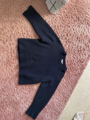 Mörkblå stickad tröja - Mysig mörkblå stickad tröja med rund halsringning och långa ärmar. Tröjan har ribbade muddar vid ärmslut och nederkant, vilket ger en klassisk och enkel look. Perfekt att matcha med jeans eller kjol för en avslappnad stil.