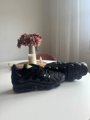 Nike Air Max Plus  Svarta Sneakers - Svarta Nike Air Max Plus TN sneakers med gulddetaljer på plös och häl. Skorna har en chunky sula med synliga luftbubblor och vågiga detaljer på ovandelen. Tillverkade i en mix av textil och syntetmaterial. Swoosh-logga i gult på sidan och dragflik bak.
