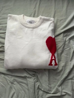 Vit stickad tröja från Ami Paris - Säljer en vit stickad tröja från Ami Paris med rund halsringning och ribbade muddar. Tröjan har en stor röd hjärtlogga med bokstaven 'A' på bröstet. Perfekt för dig som gillar stilrena plagg med en twist.