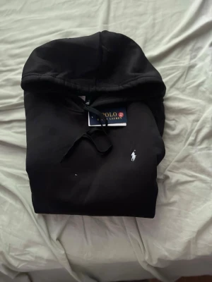 Svart hoodie från Polo Ralph Lauren - Snygg svart hoodie från Polo Ralph Lauren med klassisk huva och dragsko. Ikoniska vita loggan broderad på bröstet. Tillverkad i mjukt material som känns skönt mot huden. Perfekt för en avslappnad och stilren look.