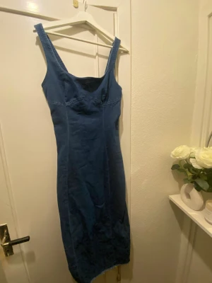 Blå jeansklänning - Snygg blå jeansklänning och figurnära passform. Klänningen har en enkel, stilren design utan synliga knappar eller dragkedja och är tillverkad i ett klassiskt denimtyg. Perfekt för dig som gillar en tidlös och cool look.