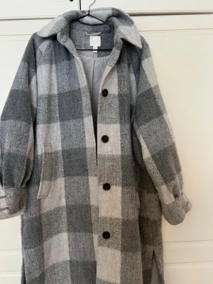 Rutig ullkappa från H&M XS - Snygg rutig kappa från H&M i grå och vita toner. Kappan har stora knappar framtill, bred krage och fickor på sidorna. Tillverkad i ullmix som ger en mjuk och varm känsla. Perfekt för kyliga dagar och trendig look. Oversized 