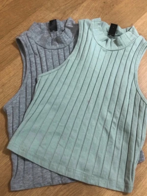 Ribbad ärmlös topp i mint och grå - Två snygga ribbade toppar i mintgrönt och ljusgrått. Modellen är ärmlös med hög hals och croppad passform, perfekta till jeans eller kjol. Materialet känns mjukt och stretchigt, troligen bomull eller polyesterblandning.den gröna har en liten sminkfläck som knappt syns, den gråa gör 75kr och den gröna för 50kr pga fläcken ifall du köper båda så är de för 100kr. Använda bara 1 gång