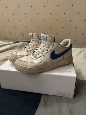 Nike Air Force 1 vita/blå sneakers - Nike Air Force 1 sneakers i vitt läder med blå swoosh och detaljer. Klassisk låg modell med perforerad tå, vit sula och snörning. Ikonisk streetstil som funkar till det mesta och ger en retrovibe. Perfekt för dig som gillar tidlösa sneakers med färgklick.