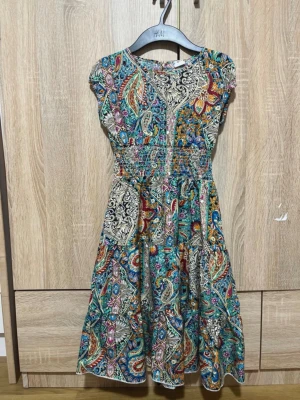 Färgglad kortklänning med paisleymönster - Säljer en färgglad kortklänning med paisley- och blommönster i blått, orange, grönt och beige. Klänningen har korta ärmar, markerad midja med smock och rund halsringning. Perfekt för dig som gillar boho-stil och vill sticka ut. Passar 11-13 år
