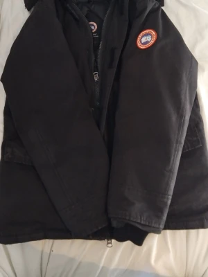 Svart dunjacka från Canada Goose - Säljer en svart dunjacka från Canada Goose med klassisk patch på ärmen och dragkedja framtill. Jackan har huva och praktiska fickor med lock. Perfekt för kalla dagar och riktigt snygg streetstil.