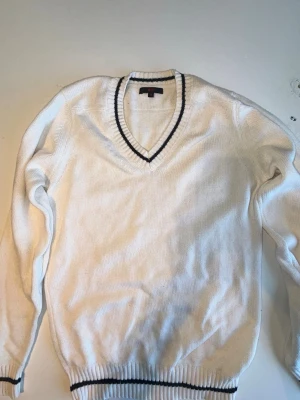 Vit stickad tröja med V-ringning BZR - Klassisk vit stickad tröja från BZR med V-ringning och svarta ränder vid hals, ärmslut och nederkant. Tröjan har lång ärm och ribbstickade detaljer som ger en sportig och stilren look. Perfekt att matcha med jeans eller kjol.