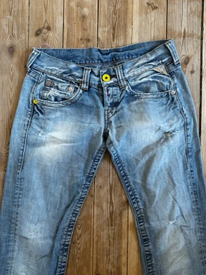 Ljusblå slitna jeans från Replay - Säljer ett par ljusblå jeans från Replay med slitningar och detaljerade sömmar. Jeansen har fem fickor, gult knappfäste och en rosa patch med blommor bak. Materialet är denim och passformen är rak. Perfekta för en avslappnad och trendig look. Otydligt vilken storlek men passar M 