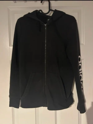 Svart Adidas hoodie med dragkedja - Säljer en svart hoodie från Adidas med dragkedja framtill och huva. Tröjan har två praktiska fickor samt Adidas-loggan i vitt längs ena ärmen som snygg detalj.(ifrån Addidas) 