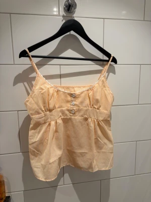 Gulvit randig linnetopp med spets - Söt gulvit randig linnetopp med tunna axelband och dekorativa knappar framtill. Toppen har en liten spetskant vid urringningen och är lätt rynkad under bysten för en luftig känsla. Perfekt för varma dagar och enkel att styla.