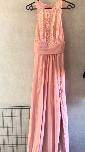 Ljusrosa långklänning med spets - Långklänning i ljusrosa med spetsdetaljer upptill och slits på kjolen. Ärmlös modell med rund halsringning och markerad midja. Perfekt för student eller bal. Kjolen är i ett mjukt, flytande material. Har använts en gång på ett bröllop. 