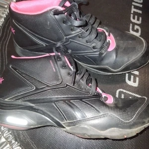 Svarta och rosa Reebok sneakers - Snygga svarta sneakers från Reebok med rosa detaljer och innersula. Skorna har högt skaft, klassisk snörning och en sportig design med diskreta mönster på sidorna. Tillverkade i syntetmaterial och textil för en cool och bekväm look.