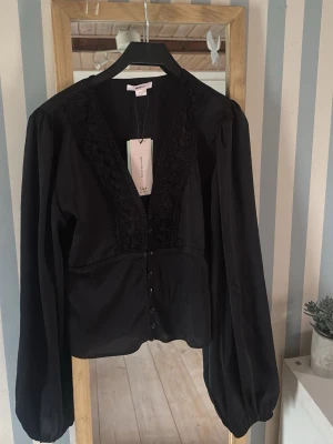Svart blus med spets från Gina Tricot - Stilren svart blus från Gina Tricot med djup V-ringning och dekorativ spets längs urringningen. Blusen har knappar framtill och långa puffiga ärmar med resår vid ärmslut. Perfekt för en trendig och elegant look. Aldrig använd, med prislapp kvar. 