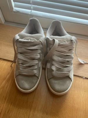 Beige Adidas sneakers i mocka - Snygga beige sneakers från Adidas med klassiska vita ränder på sidan. Skorna är låga och har en ovandel i mjuk mocka, samt breda vita skosnören och vit sula. Perfekta för en clean och stilren look. Har självklart spår på användning 