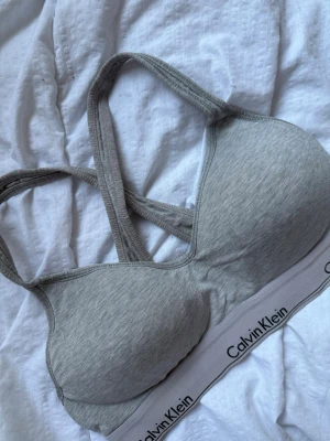 Grå sport-bh från Calvin Klein - Grå sport-bh från Calvin Klein storlek L men passar M. med vadderade kupor och bred resår med logga. Bh:n har korsade axelband i ryggen och är tillverkad i mjukt bomullsmaterial för extra komfort.