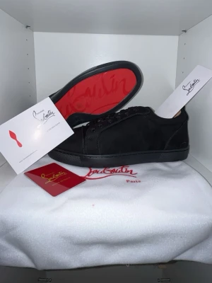 Svarta Louboutin sneakers med röd sula - Snygga svarta sneakers från Christian Louboutin i mocka med den ikoniska röda sulan. Klassisk låg modell med svarta skosnören och diskret logga på sulan. Perfekta för dig som vill ha en clean och exklusiv look.