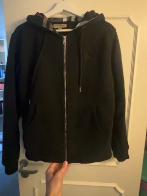 Svart zip hoodie från Burberry - Snygg svart hoodie med dragkedja från Burberry. Klassisk huva med det ikoniska rutmönstret på insidan och justerbara snören. Stora fickor framtill och ribbade muddar. Perfekt för en chill och stilren look.