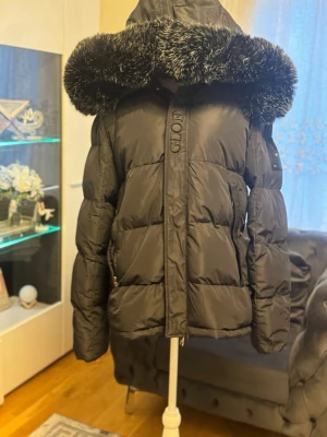 Lyxig Vinterjacka från Glorious Gangsta – Storlek L - Säljer nu en extremt snygg och varm pufferjacka från det populära märket Glorious Gangsta. Jackan är i nyskick och perfekt för kalla vinterdagar. Egenskaper: • Design: Modern och stilren i djupsvart färg med märkets exklusiva logotyp broderad framtill och ett stort, snyggt tryck på ryggen. • Huva: Stor och mysig huva med en högkvalitativ fuskpäls (faux fur) som ger en lyxig look och extra skydd mot vinden. • Passform: Storlek L (Herr), sitter riktigt bra och är mycket bekväm. • Detaljer: Rejäla 