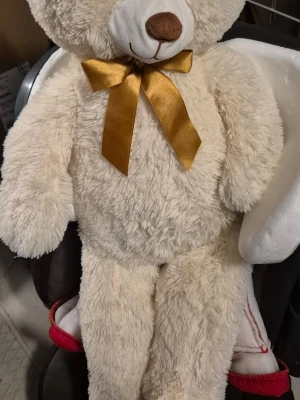 Stor mjuk nallebjörn med rosett - Säljer en stor, fluffig nallebjörn i krämvit plysch med en glansig guldfärgad rosett runt halsen. Nallen har brunt nosparti och vita detaljer på ansiktet. Perfekt som present eller mysig inredningsdetalj.