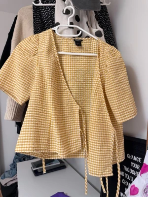 Gul och vit rutig omlottopp Lindex - Söt gul och vit rutig omlottopp från Lindex med korta ärmar och knytband i midjan. Toppen har en avslappnad passform och är gjord i ett lätt bomullsmaterial, perfekt för varma dagar.