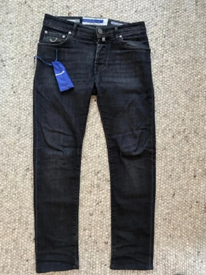 Jacob Cohen Nick slim jeans strl 30 - Snygga svarta jeans från Jacob Cohen med klassisk femficksmodell och silverfärgade detaljer. Jeansen har en diskret tvättad look och är tillverkade i stretchigt denim för extra komfort. Perfekta för dig som gillar en smal passform och stilren design. Hör av er vid frågor och funderingar! 