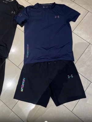 Under Armour träningsset herr - Säljer ett träningsset från Under Armour med t-shirt och shorts. Finns i marinblått och svart. Setet har logga och text i regnbågsfärger, tillverkat i snabbtorkande funktionsmaterial. Perfekt för gym eller löpning. Finns i storlek S och M inne. Bara att höra av sig 