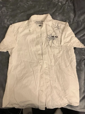 Vit kortärmad skjorta Rare Flux - Vit kortärmad skjorta från Rare Flux med diskreta ränder och blommigt tryck på bröstet. Skjortan har klassisk krage och knappar framtill. Perfekt för dig som gillar en clean men ändå unik stil.