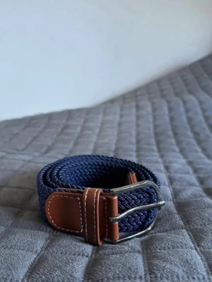 Mörkblått flätat bälte med skinn 105 cm - Snyggt mörkblått flätat bälte med fina detaljer i brunt skinn och ett stilrent silverfärgat spänne. Det har en klassisk look som passar lika bra till jeans som till chinos. Den flätade designen gör att du enkelt kan justera längden så att det sitter perfekt. Längd: 105 cm.