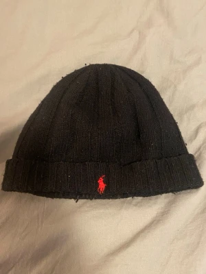 Svart mössa från Polo Ralph Lauren - Säljer en svart ribbstickad mössa från Polo Ralph Lauren med den klassiska röda loggan broderad framtill. Mössan är tillverkad i 80% lammull och 20% nylon, vilket gör den både varm och mjuk. Perfekt accessoar för kyliga dagar. Den luddar av sig ganska mycket dock men ändå ricktig stilig.