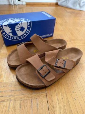 Bruna Birkenstock sandaler mocka - Säljer ett par klassiska bruna Birkenstock sandaler i mocka med två breda remmar och justerbara metallspännen. Fotbädden är ergonomiskt utformad för extra komfort och sulan är i kork. Perfekta för dig som gillar stilren och avslappnad look. Aldrig använda då dem ej passa mig! Storlek 45. Hör av er vid frågor☺️