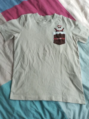 Vit t-shirt med julficka och tomte - Rolig vit t-shirt med tryck av en tomte som sticker upp ur en ficka designad som en skorsten. På fickan står det 'I Sleigh!'. Perfekt för dig som vill ha en unik och skön t-shirt till julen. Tillverkad i mjuk bomull och har korta ärmar.