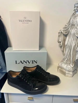 Svarta sneakers från Lanvin - Stilrena svarta sneakers från Lanvin med blank tå och detaljer i både läder och mocka. Skorna har svarta skosnören och en klassisk låg siluett. Perfekta för dig som gillar en clean och lyxig look.