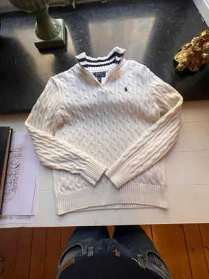 Vit kabelstickad tröja Polo Ralph Lauren - Vit kabelstickad tröja från Polo Ralph Lauren med halv dragkedja och ribbade muddar. Tröjan har en liten mörkblå logga broderad på bröstet och en hög krage med randig insida. Perfekt för lager på lager. Perfekt skick(använd fåtal gånger varsamt, inga defekter) 14-16 år passar bra ifall du är 160-165. Nypris: 1299kr 