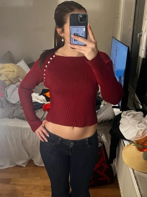 Vinröd ribbad croptröja med knappar - Säljer en vinröd ribbad croptröja med långa ärmar och dekorativa vita knappar på axeln. Tröjan har en tight passform och är tillverkad i ett stretchigt material som sitter snyggt på kroppen. Perfekt för dig som gillar trendiga och stilrena plagg. Liknar den trendiga Zara toppen