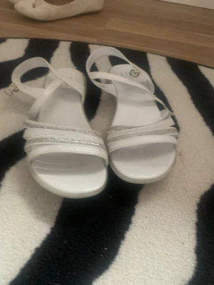 Vita sandaler med glitterdetaljer - Säljer ett par vita sandaler med smala remmar och snygga glitterdetaljer. Sandalerna har en justerbar ankelrem med spänne och öppen tå. Perfekta för sommarens alla tillfällen och ger en fräsch look till din outfit.