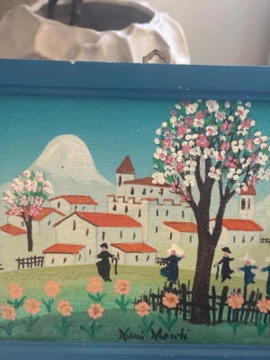 Färgglad tavla med bymotiv - Säljer en liten tavla med blå träram och motiv av en by med vita hus, röda tak, blommande träd och människor i landskapet. Signerad av konstnären Yani Monti. Perfekt som unik inredningsdetalj.