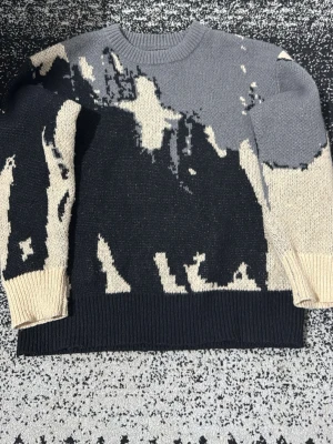 Stickad tröja i grått och beige mönster - Unik stickad tröja med abstrakt mönster i svart, grått och beige. Tröjan har rund halsringning, långa ärmar och ribbade muddar. Perfekt för dig som gillar statement-plagg och vill sticka ut lite extra.