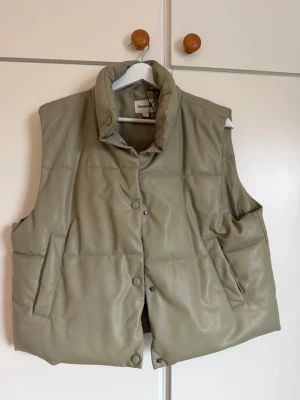 Väst från Kappahl - Stilren beige skinnväst från Kappahl i storlek S. Västen har hög krage, tryckknappar framtill och två snedställda fickor. Perfekt att slänga över en hoodie eller tröja för extra värme.