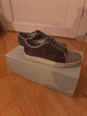 Lanvin grå sneakers i mocka och lackdetaljer - Snygga grå sneakers med ovandel i mocka och glansiga lackdetaljer på tå och runt kanten. Skorna har vita sulor och matchande grå skosnören. Perfekta för dig som gillar stilrena och moderna sneakers med en twist. Uk 5 men sitter som en 41 på foten (stora i storleken) Låda, två dustbags och extra skosnören följer med. Vid snabb affär kan priset diskuteras.