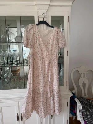 Blommig omlottklänning från Newbie - Söt långklänning från Newbie i vitt med små rosa blommor. Klänningen har omlottmodell med knyt i midjan, puffiga korta ärmar och volang nedtill. Perfekt för sommarens alla tillfällen!