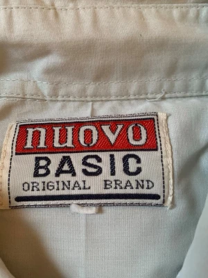 Vit skjorta från Nuovo Basic - Klassisk vit skjorta från Nuovo Basic med knappar framtill och två bröstfickor. Skjortan har en stilren design med normal passform och lång ärm. Perfekt att styla till olika outfits för en clean look.