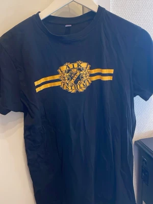 Svart AIK t-shirt med gult tryck - Snygg svart t-shirt från AIK med gult tryck och klubbens emblem på bröstet. Klassisk passform och rund hals. Perfekt för dig som vill visa ditt AIK-stöd på ett stilrent sätt. Materialet känns som bomull.