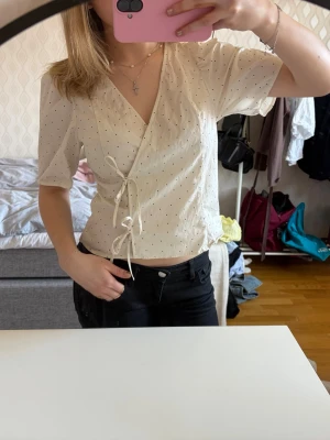 Vit omlottblus med prickar och knyt - Söt vit omlottblus med små svarta prickar och knytband i sidan. Blusen har v-ringning och korta puffärmar. Tillverkad i ett lätt och luftigt material som passar perfekt till jeans eller kjol. Enkel men trendig design som ger en fräsch look.